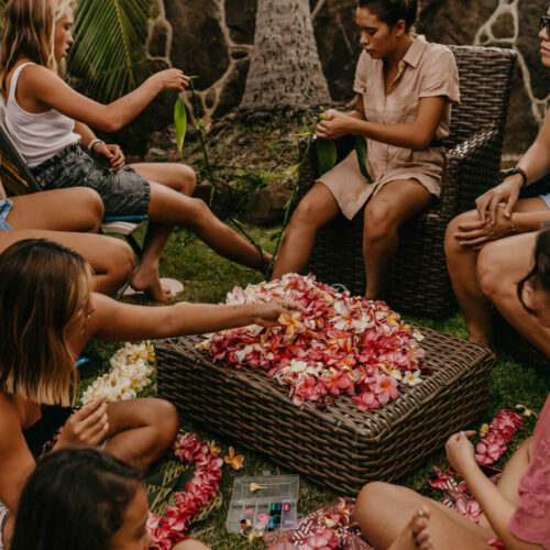 A group of people sit around a wicker table covered with colorful flower petals, stringing leis in an outdoor setting with wicker furniture.